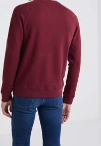 Sudadera burdeos con cuello redondo, puños y dobladillo acanalados. Presenta una textura suave y un diseño sencillo. Combinada con jeans azules.