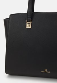 Sort struktureret tote bag med tekstur, der har et gyldent firkantet hardware-detalje og et præget logo nederst.