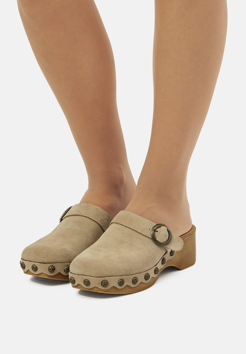 Zuecos de ante en beige con puntera redondeada, una hebilla circular decorativa y un tacón tipo madera, con un borde festoneado y acentos metálicos.