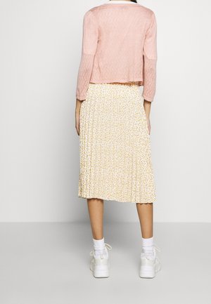 Hellrosafarbenes, cropped Sweatshirt mit einem strukturierten Muster, kombiniert mit einem weißen, plissierten Rock mit gelbem Blumenmuster. Weiße Sneaker runden den Look ab.