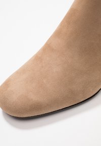 Beige mockasinhöst boots med spetsig tå, slät yta och en smal, diskret silhuett. Platt svart sula synlig nederst.