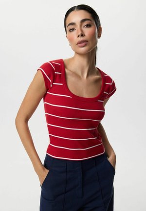 Kobieta o ciemnych włosach, ubrana w czerwoną bluzkę z krótkim rękawem z białymi paskami i granatowe spodnie, stoi z rękami w kieszeniach na białym tle.