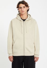 Cremfarbener Zip-Hoodie aus weichem Stoff, mit einer Vordertasche, Kordelzugkapuze und gerippten Bündchen. Minimales Logo auf der Brust.