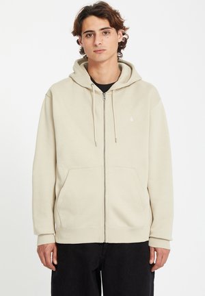Cream-colored zip-up hoodie made of soft fabric, featuring a front pocket, drawstring hood, and ribbed cuffs. Minimal logo on chest.