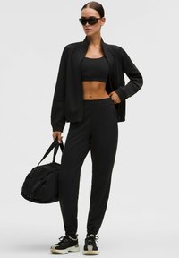 Juodas sportinis apranga, kurioje yra trumpas viršus, užtrauktuku užsegama striukė ir siaurėjantys kelnių. Blizgios juodos saulės akiniai ir juoda sportinė kuprinė baigia išvaizdą.