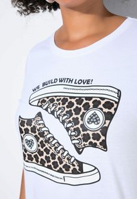 Biała koszulka z grafiką przedstawiającą tenisówki w panterkę, ozdobioną cyrkoniami, oraz napisem "BUDUJEMY Z MIŁOŚCIĄ!" powyżej.
