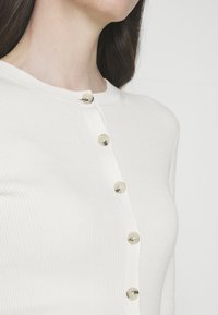 Cardigan bianco a coste con scollo rotondo e sei bottoni dorati sul davanti. La trama appare morbida e il materiale sembra aderente.