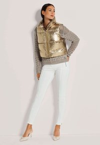 Metallisch goldene Pufferweste mit Reißverschluss, kombiniert mit einem hellgrauen Strickpullover und hellblauen, figurbetonten Hosen, abgerundet durch spitze Absätze.