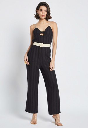 Salopette noire avec des bretelles fines, motif texturé, décolleté en forme de trou de serrure et jambes larges ; cintrée avec une ceinture crème ornée d'une boucle circulaire.