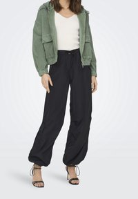 Zielona kurtka z korduroju o luźnym kroju, z dwiema przednimi kieszeniami, zestawiona z czarnymi, luźnymi spodniami na sznurek oraz czarnymi sandałkami na obcasie.