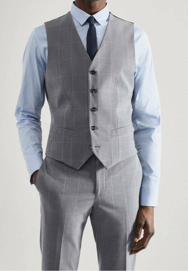Homme portant une chemise bleu clair, une cravate bleu marine, un gilet à carreaux gris et un pantalon assorti, les mains partiellement dans les poches.