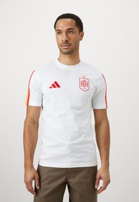 Hvit bomull t-skjorte med røde og gule detaljer på ermene, et fremtredende rødt Spania-emblem på brystet, og Adidas-logo på forsiden.