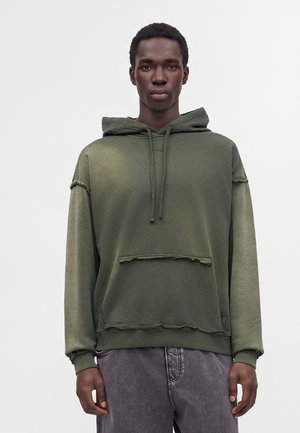 Ung mand iført en olivengrøn hættetrøje med frynset kant og grå falmede jeans, der står foran en ensartet lys baggrund.