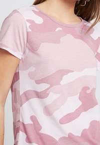 Růžové maskáčové tričko vyrobené z měkké tkaniny, má klasický kulatý výstřih, krátké rukávy a jemný, smíšený vzor v různých odstínech růžové.