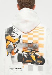 Bag-til af hvid hoodie med McLaren Formula 1 bil, jubelende teammedlemmer i orange, og ternet flagmønster.