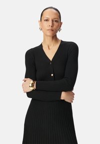 Abito nero a costine con scollatura a V, maniche lunghe e discreti bottoni dorati. Modella accessoriate con un bracciale dorato. Design semplice e aderente.