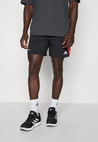 Homme portant un short de sport noir Adidas avec des panneaux latéraux rouges, des baskets Adidas noires et blanches, et des chaussettes blanches Adidas, se tenant devant un fond blanc.