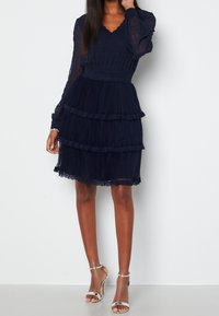 Robe de couleur marine avec un design à volants, texture plissée, accents en dentelle, et manches longues. Portée avec des sandales à talons argentées.