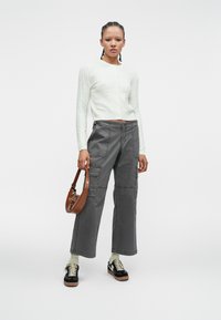 Biely kardigán s pleteným vzorom, šedé cargo nohavice s vreckami, hnedá kabelka cez plece a čierne tenisky s ľahkými podrážkami. Pevné farby a textúry.