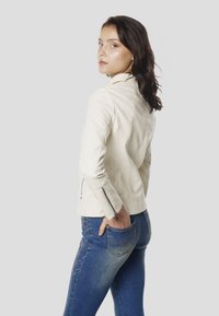 Krémová kožená bunda s priliehavým strihom, diagonálnymi švami a zapínaním na zips na manžetách, v kombinácii s modrými rifľami s vyšívanými detailmi.