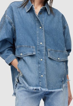 Personne portant une veste en jean bleu clair oversize avec boutons-pression, poches à rabat, ourlet brut, et un jean déchiré assorti.