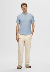 Svetlo modra kratko rokavna polo majica z diskreto rebrasto strukturo, v kombinaciji s smetanastimi nabranimi hlačami in rjavimi sandalami, roke v žepih.