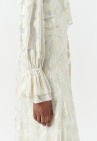 Valkoinen läpikuultava kangas, jossa on kultaisia pyöreitä kuvioita, varustettuna röyhelöhihoilla ja joustinhihalla. Siluetti on virtaviivainen ja siinä on teksturoitu pinta.