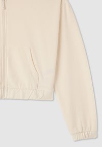 Giacca cropped con zip in tessuto beige chiaro. Presenta una tasca a marsupio e polsini elasticizzati. Texture liscia con dettagli minimi.