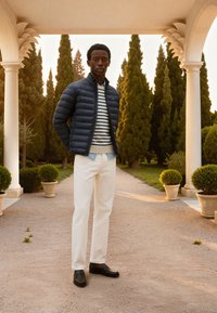 Un jeune homme portant une doudoune bleu marine et un pull à rayures se tient sous une colonnade avec de grands arbres à feuilles persistantes en arrière-plan.