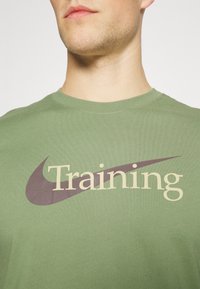Camiseta de algodón verde oliva con un gran gráfico de "Entrenamiento" en crema y púrpura oscuro, con un cuello clásico y mangas cortas.