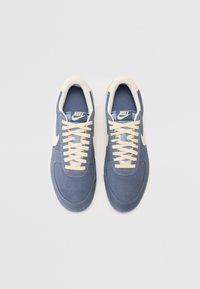 Modré semišové tenisky s krémovými šňůrkami a akcenty, s bílým logem Nike a prošíváním. Plochá podrážka a design s kulatou špičkou.