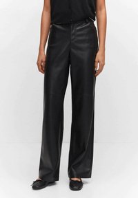 Femme portant un pantalon en cuir noir taille haute à jambes larges, associé à des ballerines noires, debout devant un fond blanc uni.