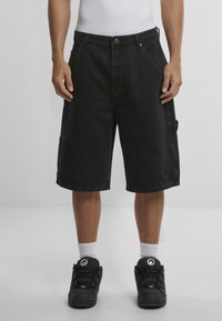 Pantalones cortos de denim negro con un corte holgado, bolsillos y cierre de botón. Acabados con un dobladillo limpio y combinados con zapatillas negras y calcetines blancos.