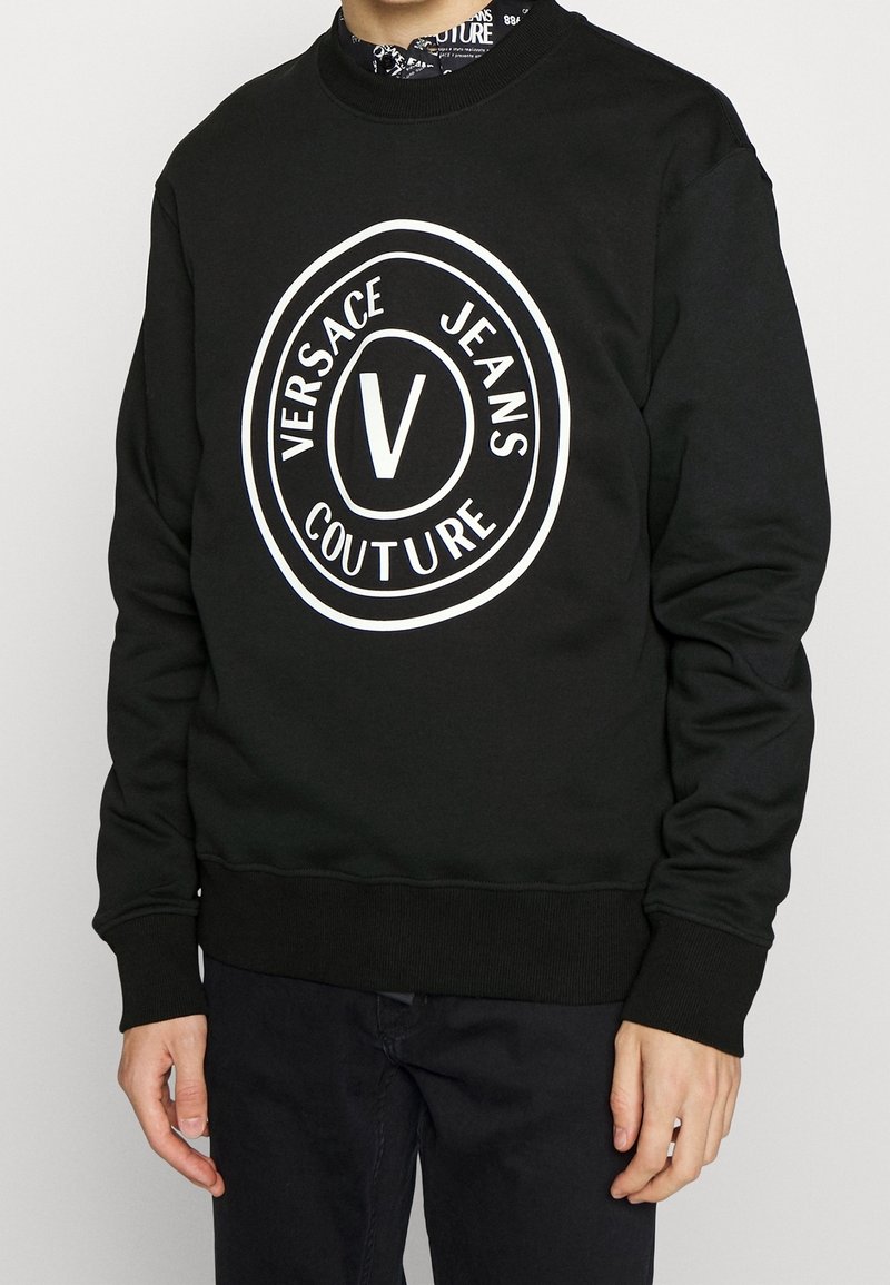 Svart sweatshirt med en stor vit cirkulär logotyp med texten "VERSACE JEANS COUTURE" och ett "V" i mitten. Ribbstickade muddar och fåll.