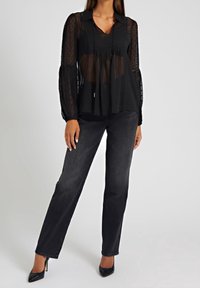Blouse noire transparente à motif pois, à boutons sur le devant, avec manches longues bouffantes. Associée à un jean droit foncé et des talons hauts noirs.