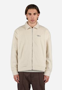 Cream-colored fleece jacket with a zip closure, pointed collar, and embroidered logo. Features a soft texture and relaxed fit.