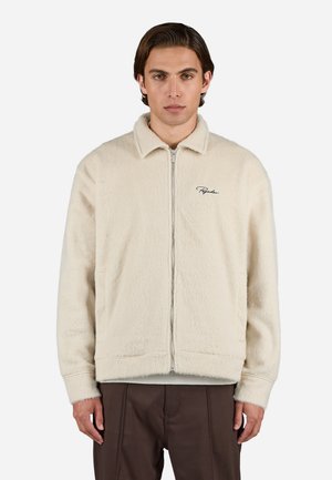 Cream-colored fleece jacket with a zip closure, pointed collar, and embroidered logo. Features a soft texture and relaxed fit.