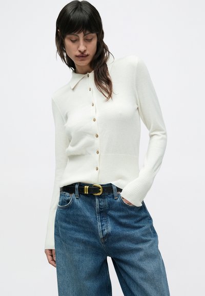 Λευκό πουλόβερ με κουμπιά και γιακά, με χρυσά κουμπιά, εφαρμοστή γραμμή και ριμπ τελείωμα. Συνδυάζεται με μπλε ψηλόμεσα τζιν και μαύρη ζώνη.