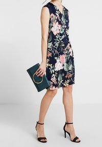 Sukienka w kwiatowy wzór w kolorze granatowym z krótkimi rękawami i marszczonym detalem. Połączona z zieloną torebką kopertową z okrągłym uchwytem i czarnymi sandałami na obcasie.
