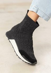 Botte chevillère tricotée noire avec motif texturé, dotée d'une semelle intermédiaire blanche et d'une semelle extérieure en caoutchouc noir. Le design inclut un col élastique pour un ajustement confortable.