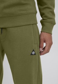 Olivengrüne Sweatpants aus weichem Stoff, mit einem gerippten Bund und einem dreieckigen schwarzen Logo-Patch an der Seitentasche.