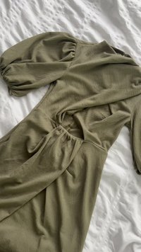 Olivengrønn kjole med puffede ermer og innsnevret midjedetalj, lagt flatt på et hvitt teksturert sengeteppe.