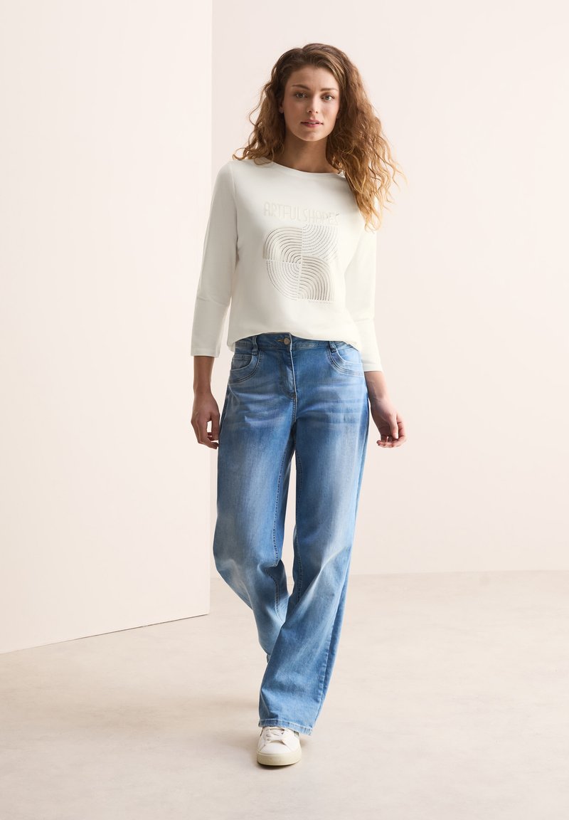 Weißes langärmliges Sweatshirt mit dem Aufdruck "KÜNSTLERISCHE FORMEN", kombiniert mit hellblauen weit geschnittenen Jeans und weißen Sneakers. Sanfter Stoff und lässiger Stil.