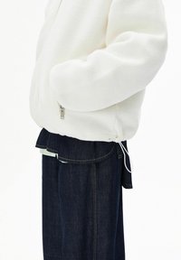 Weiße Fleecejacke mit Reißverschluss, Seitentaschen und verstellbarem Kordelzug am Saum, kombiniert mit dunkelblauen Jeanshosen mit einem Lagen-Saum.