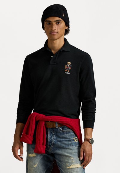Polo noir à manches longues avec logo d'ours brodé, associé à un jean déchiré et un pull en maille rouge noué autour de la taille.
