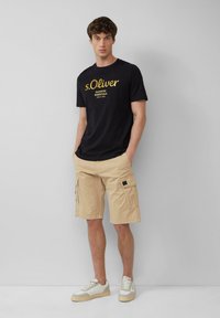 Schwarzes Grafik-T-Shirt mit goldener Schrift, beigefarbene Cargo-Shorts mit Seitentaschen und beige Sneakers mit weißen Akzenten. Einfarbig grauer Hintergrund.