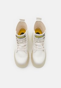 White leather ankle boots with a textured finish, lace-up design, and clear rubber soles. Features a yellow and black logo label on the tongue.