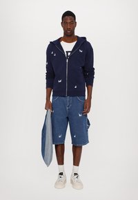Mornarska zip-up hoodie s bijelim vezom zečice, uparen s plavim trapericama kratkim hlačama koje imaju sličan vez. Model drži traper jaknu.