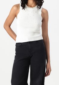 Femme portant un top blanc sans manches en maille côtelée avec des détails en perles et perles au niveau du col et un pantalon noir taille haute sur fond blanc.