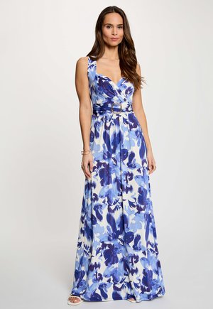 Mujer con cabello largo y castaño que lleva un vestido maxi sin mangas azul y blanco con flores y un cinturón con hebilla cuadrada, de pie frente a un fondo liso.